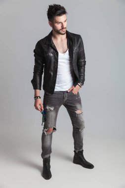 Güneş gözlüklerini tutan deri ceketli seksi genç adamı yan cebinde, bir eliyle hafif gri arka plan üzerinde dururken görünüyor