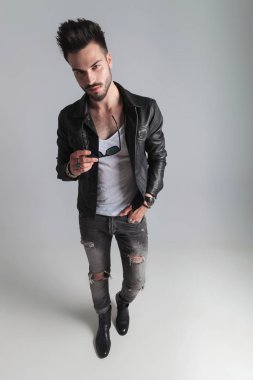 deri ceket ve gri yırtık kot tutan güneş gözlüğü eli cebinde, geniş açı resim ile ışık gri arka plan üzerinde duran adam