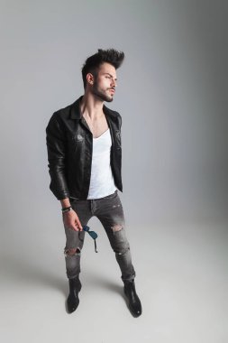 deri ceketli cool adam güneş gözlüğü tutar ve açık gri renkli, tam uzunlukta resim üzerinde dururken yan görünüyor