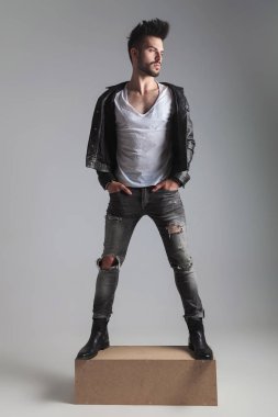 elleri ceplerinde ahşap sandık üzerinde duran seksi erkek tarafına ışık gri arka plan üzerinde görünüyor. O bir deri ceket ve gri kot, tam vücut resim giyiyor
