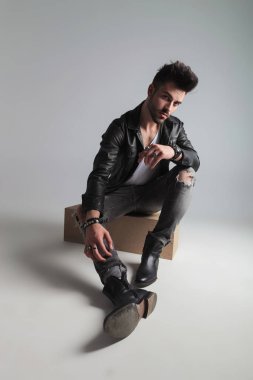 bir siyah deri ceket giymiş seksi adam açık gri renkli bir ahşap sandık oturur ve tarafa eğilir