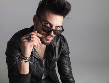 portrait of seductive man wearing leather jacket looking over sunglasses while sitting on light grey background and smiling clipart