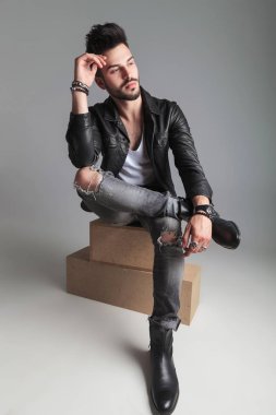 tarafa, açık gri renkli deri ceket ve bağdaş kurup oturmuş bir ahşap kutu üzerinde gri yırtık kot dalgın adam görünüyor