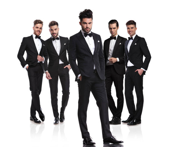 группа из пяти красивых женихов с расслабленным лидером, стоящим перед ними с руками в карманах, на белом фоне
