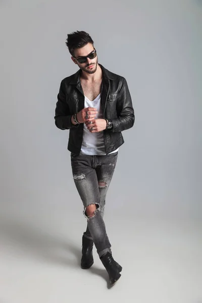 man wearing leather jacket and gray jeans looks to side while standing on white background with legs crossed and fixing his rings, full length picture