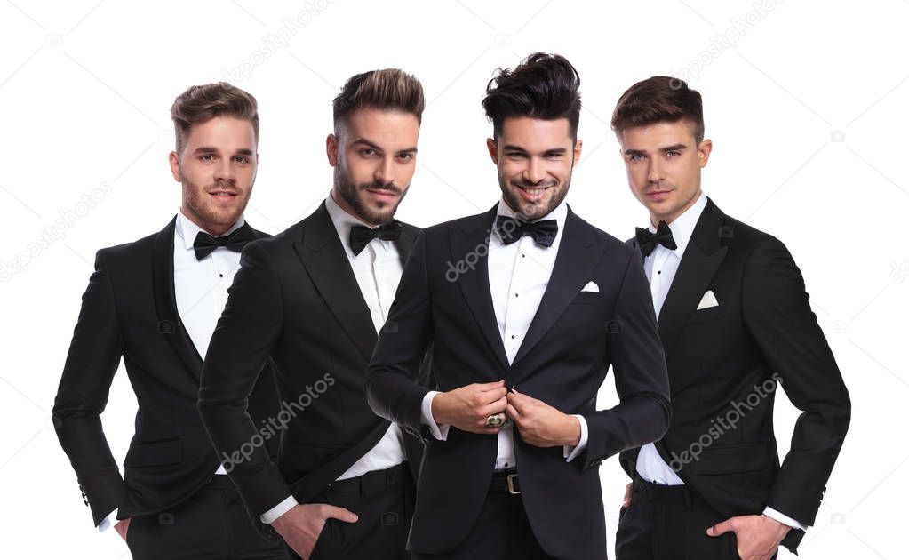 four young men in black tuxedoes standing together on white background with hands in pockets, one of them buttoning his suit, portrait picture