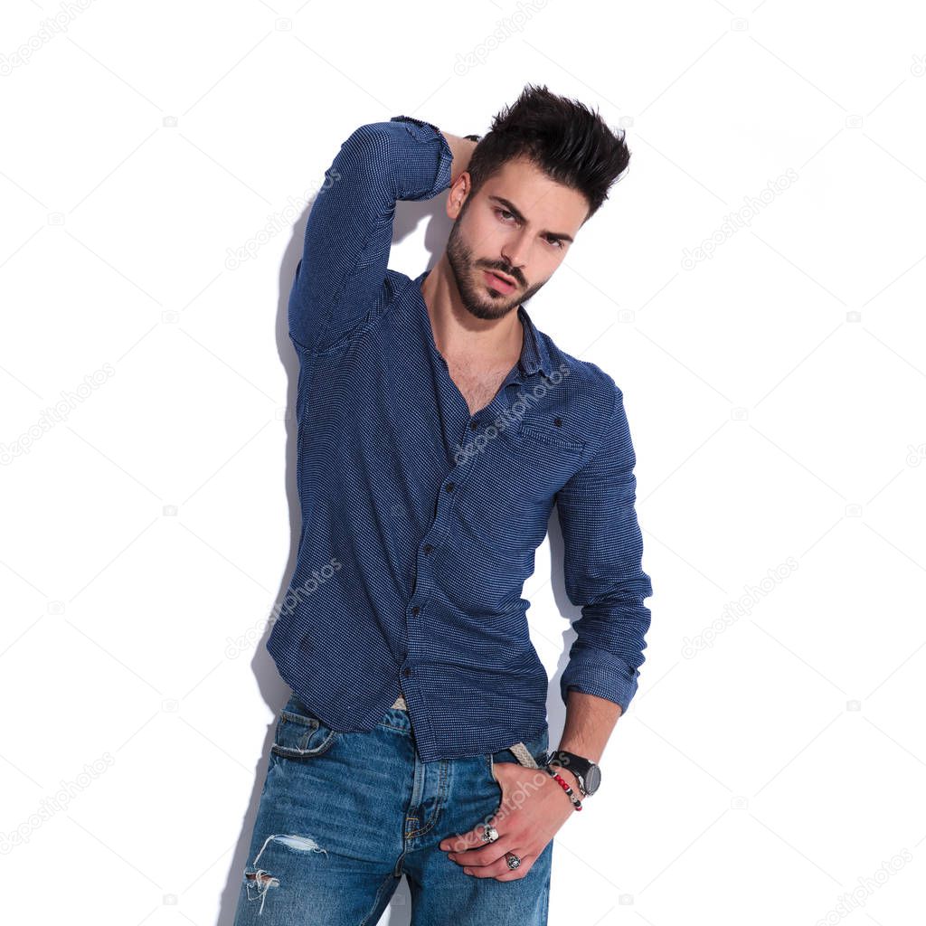 portrait sensual man wearing a navy shirt posing with a hand in pocket and the other his backhead while standing near a white wall