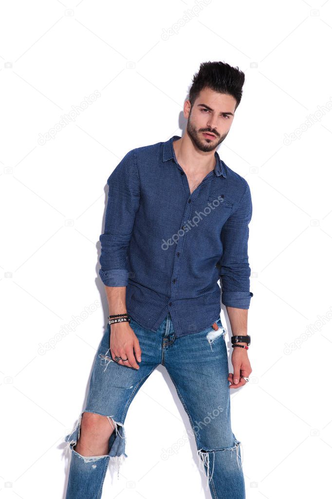 attractive man wearing a partly undone navy shirt and ripped jeans standing near a white wall, portrait picture