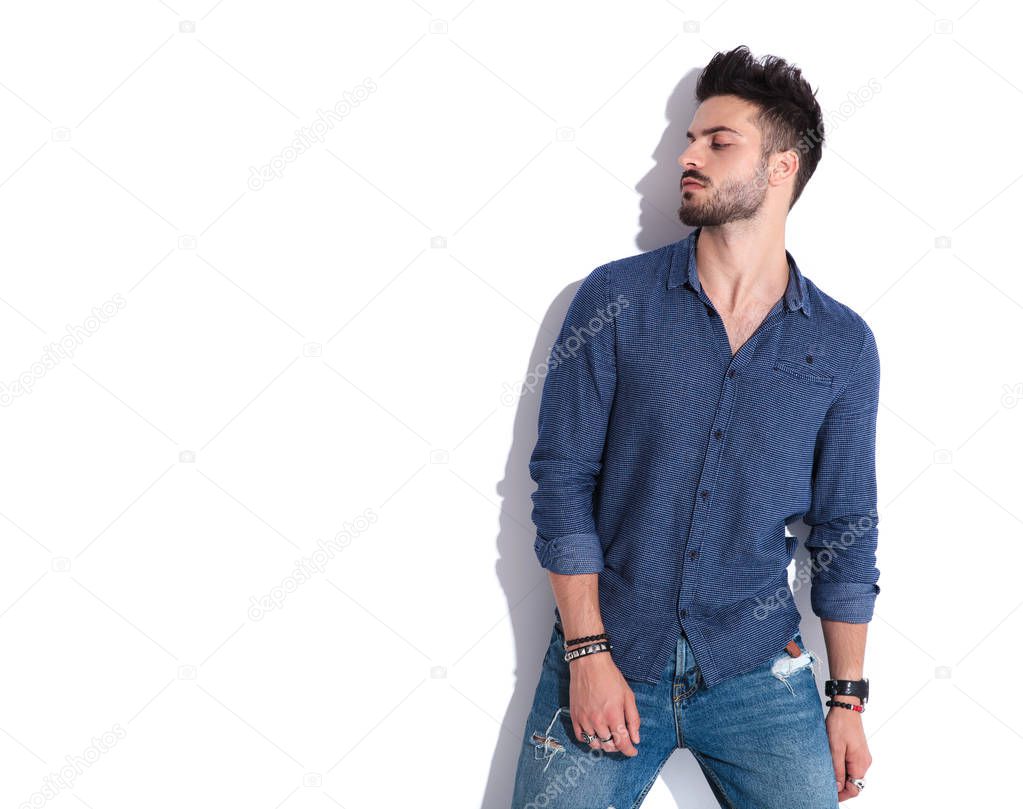 portrait of young casual man wearing a Guess shirt looking down to side while standing near a white wall