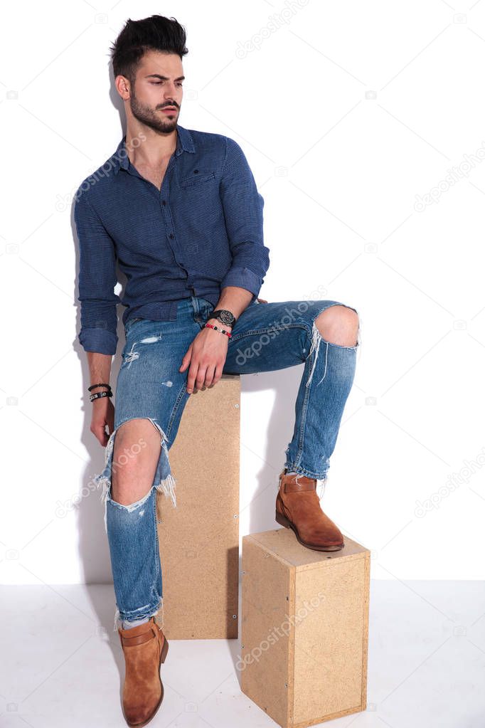 man wearing a navy shirt sitting on wooden box and looking down to side while resting his foot on another wooden box, full length picture
