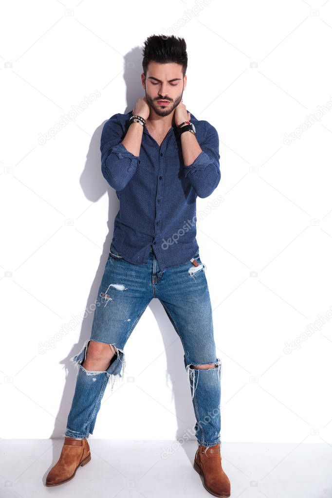 man in navy shirt and ripped jeans posing while holding his neck with both hands. and looking down. He is standing near a white wall, full body picture