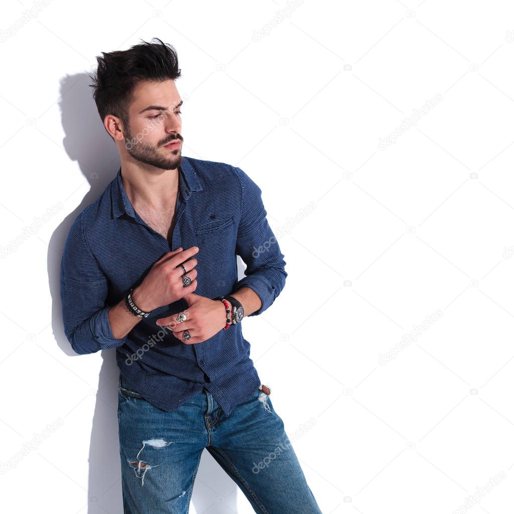 attractive man in navy shirt and blue jeans looking down to side while standing near a white wall, fixing his rings, portrait picture