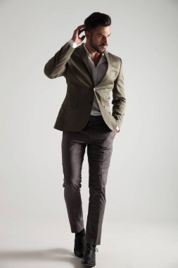 Yeşil elbiseli adam cebinde el ile ışık gri arka plan üzerinde saçı düzenlenmesi ve tarafına bakarak yürüyor