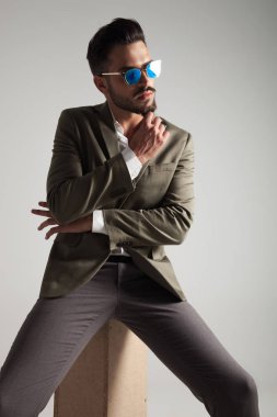 Yeşil elbiseli şık adam düşünüyor ve açık gri renkli bir ahşap sandık üzerinde otururken yan görünüyor