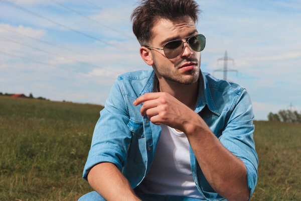 portrait of handsome casual man wearing sunglasses and denim shirt crouching in a field outside and looking to side