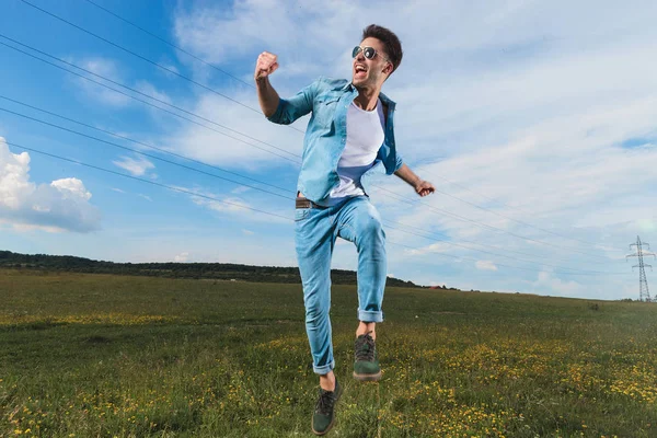 カジュアル デニム シャツとサングラス外フィールドでジャンプと空中で拳を祝う男を興奮 — ストック写真