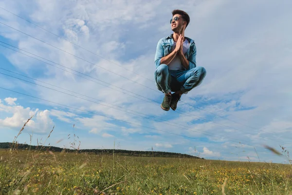 芝生のフィールドでジャンプと側に見ながら祈っているサングラスをかけたカジュアルな男 — ストック写真