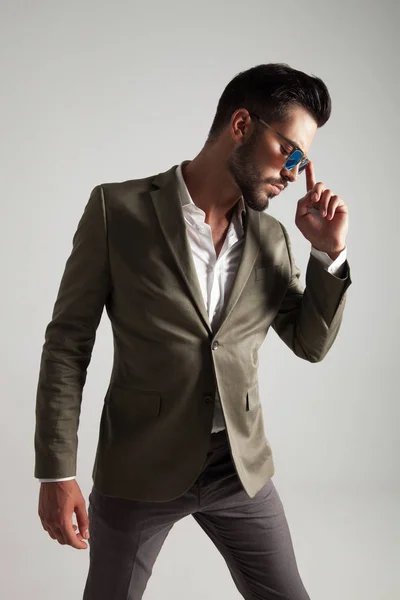 pensive man with green suit and sunglasses looks down to side whlie standing on light grey background, fixing his sunglasses