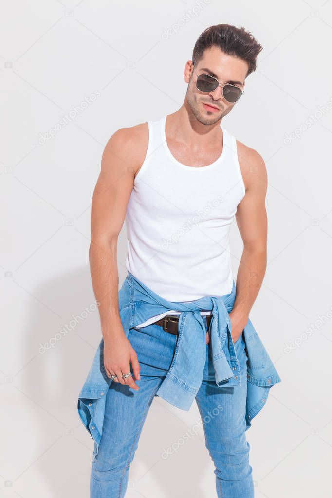 portrait of seductive casual man with denim shirt around waist standing with a hand in pocket in spotlight near a white wall. He is wearing a pair of sunglasses and jeans