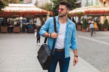 yakışıklı genç adam bir sokakta şehirde ayakta tarafına aşağıya bakar. O bir çift güneş gözlüğü giyiyor ve omuz çantası