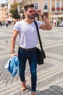 onun güneş gözlüğü ve görünüyor sabitleme şehirde yürüyen casual adam mavi gömleğini tutarak yan tam vücut resim