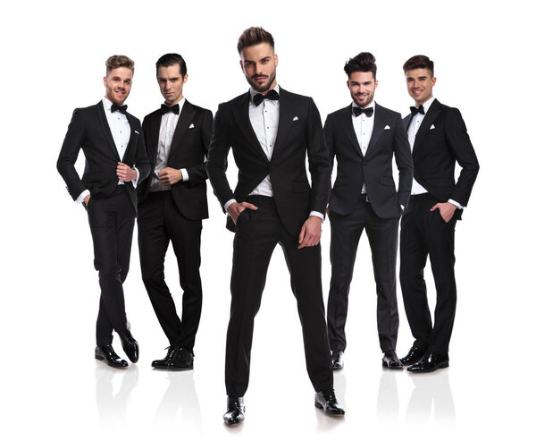 five elegant grooms in black tuxedoes with relaxed leader standing in front on white background, with hands in pockets