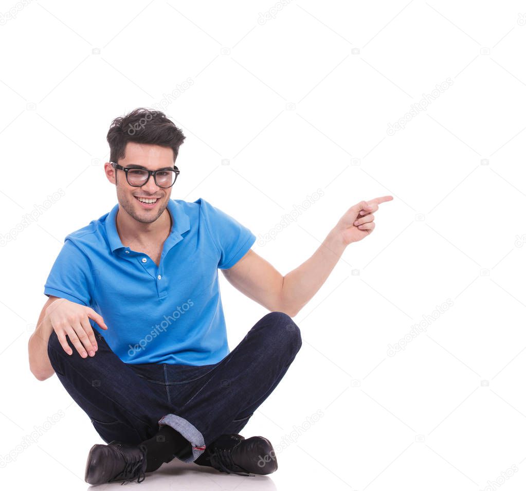 young smiling casual man sitting and pointing to side on white background