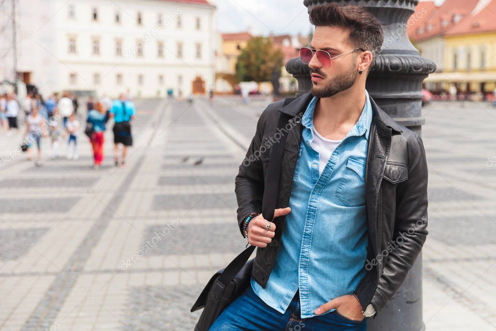 portrait of handsome casual man in the city looking to side while leaning on a lighting pole. he is wearing a leather jacket and a pair of sunglasses