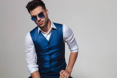 ışık gri arka plan üzerinde otururken bir mavi yelek ve güneş gözlüğü takmış seksi adam portresi