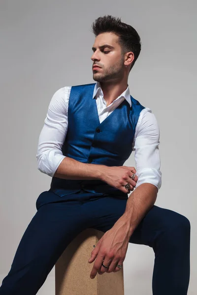 portrait of sexy seated man in blue vest looking down to side while fixing his sleeve on light grey background