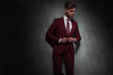 portrait of relaxed handsome man buttoning his red suit and looking to side while leaning against a grey wall clipart