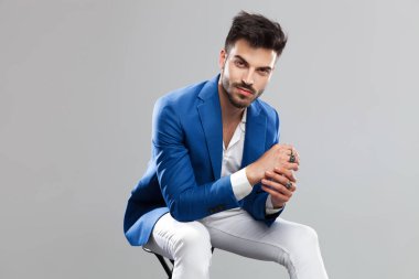 ahşap bir taburede oturan ve avuç içi bir arada hafif gri arka plan üzerinde tutan gülümseyen smart casual adam portresi