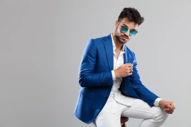 ışık gri arka plan üzerinde otururken dizleri üzerinde dirseğini dinlenme güneş gözlüğü takıyor akıllı casual adam portresi