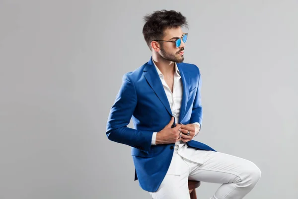 portrait of handsome seated man with sunglasses buttonning suit and looking to side on light grey background