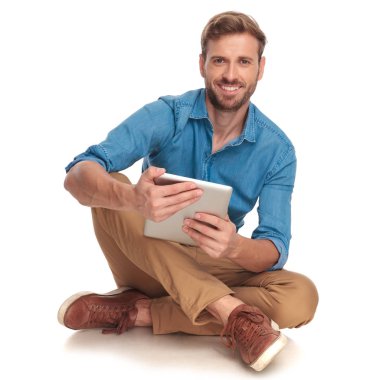 Beyaz arka plan üzerinde oturan ve tablet tutan mutlu casual adam gülümsüyor