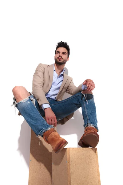 casual man in brown suit resting on wooden boxes and looking to side on white background
