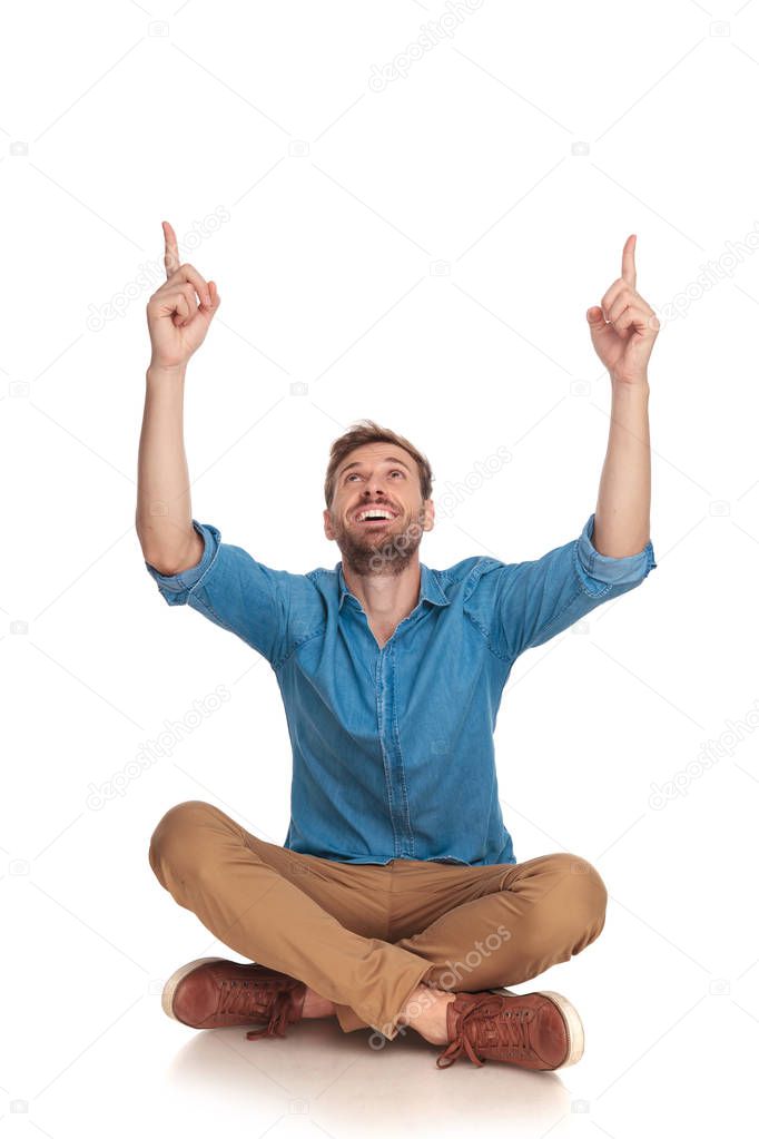 young smiling casual man is sitting and pointing up at something on white background