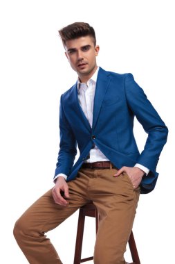 Beyaz arka plan üzerinde cebinde tek elle bir tabure üzerinde oturan rahat genç adam