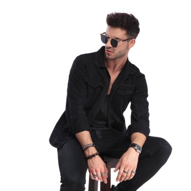 siyah elbise ve beyaz arka plan üzerinde tarafına bakarak güneş gözlüğü takan oturmuş rahat adam portresi