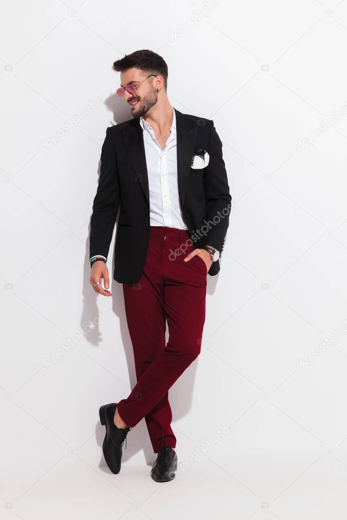 attractive smart casual man looks to side and smiles while leaning against a white wall, looking relaxed, full body picture