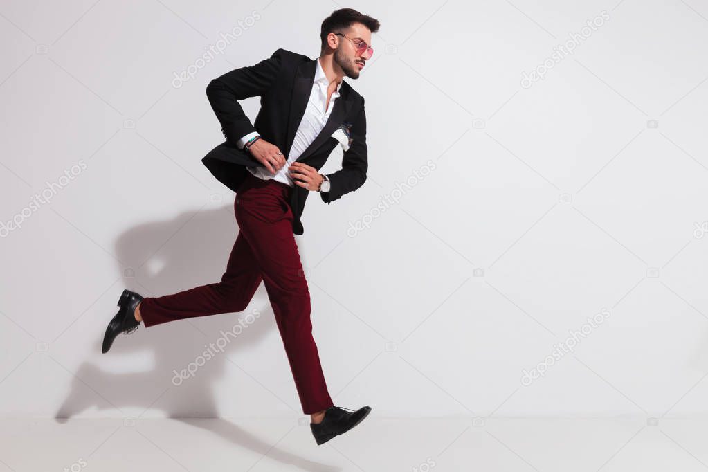 side view of elegant man with red sungless running near a white wall and buttoning his black suit, full length picture