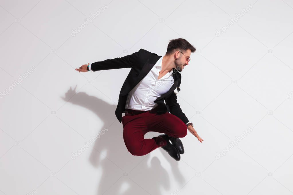 smart casual man jumping and doing a heel click near a white wall while looking down to side, full body picture
