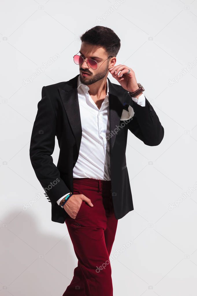 portrait of stylish young man wearing black suit and red sunglasses stepping to side and looking behind on white background