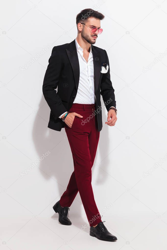 elegant man wearing undone black suit and yellow sunglasses walking to side with a hand in pocket on white background, full length picture