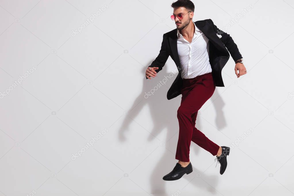 elegant young man wearing black suit and red sunglasses leaping and looking to side on white background