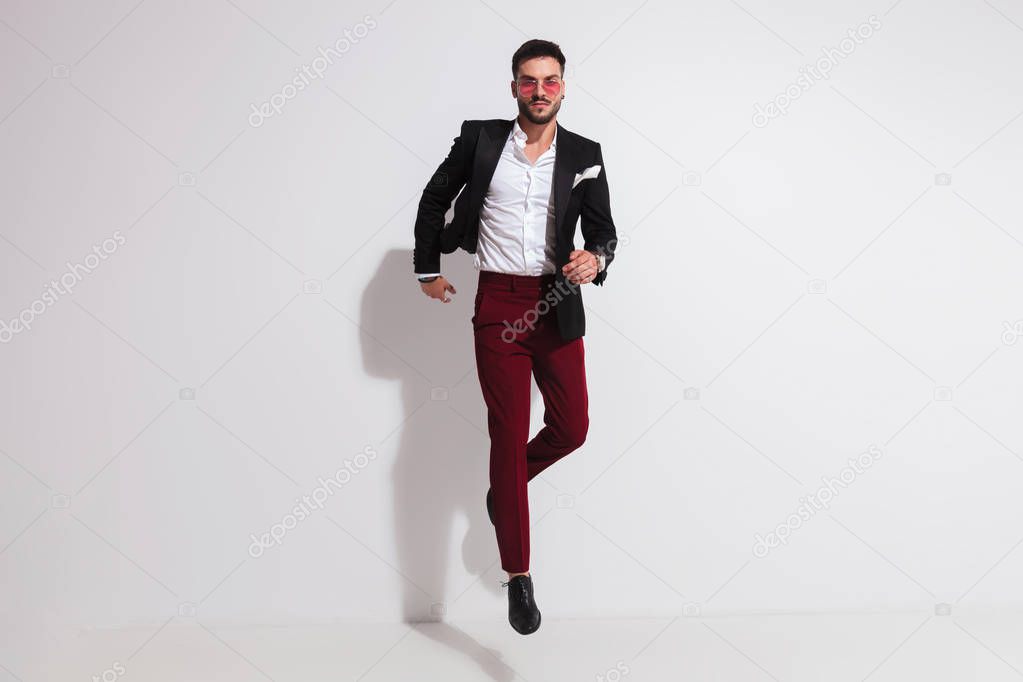 elegant man wearing a black suit and red sunglasses jumping in the air and posing like running near a white wall