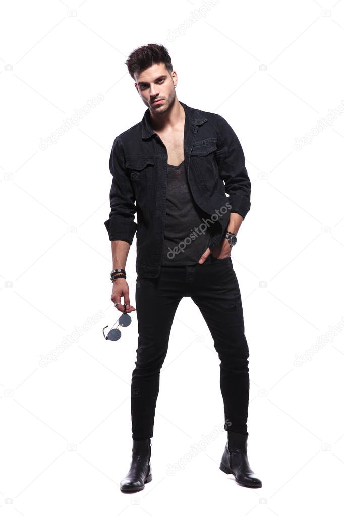 relaxed young man in black clothes standing on white background and holding sunglasses, looking cool and macho, full body picture