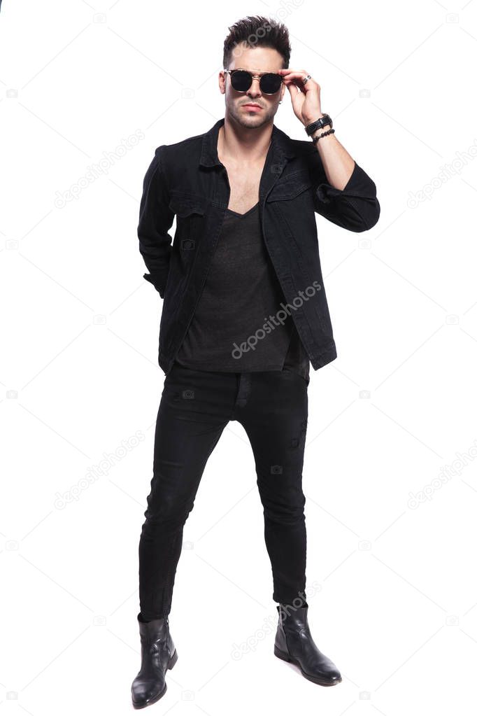 cool young man in black clothes arranges sunglasses while standing on white background, full length picture