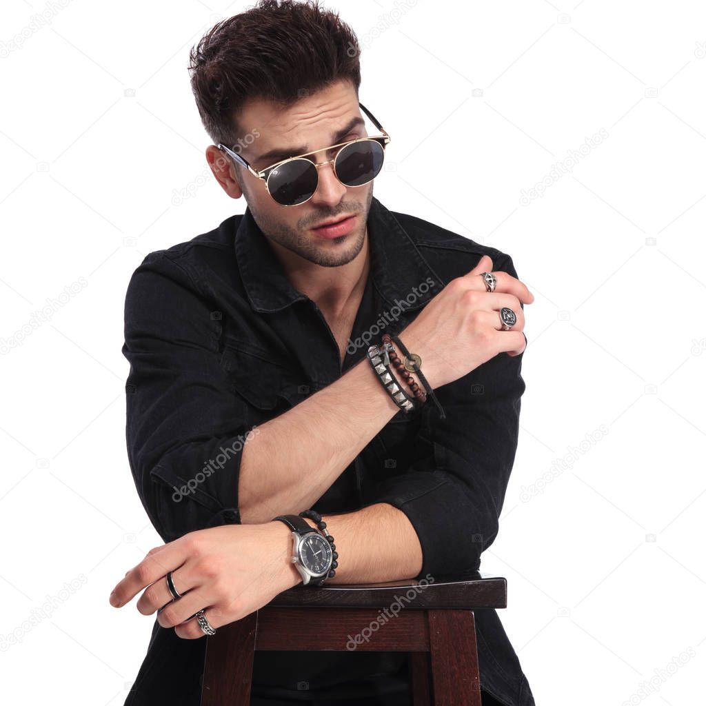 portrait of surprised young man wearing black clothes and sunglasses leaning on wooden table and looking down to side on white background