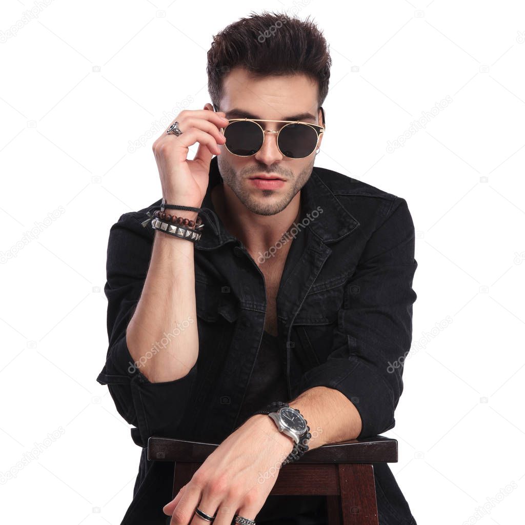 portrait of young man in black casual clothes leaning on wooden chair and arranging his sunglasses while standing on white background